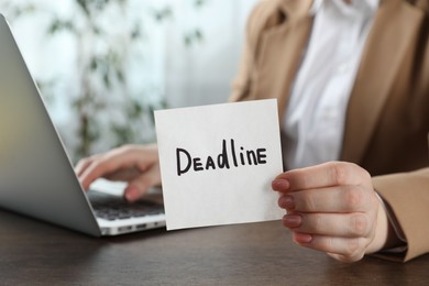 Photo of Woman holding sticky note with word Deadline at wooden table in office, closeup