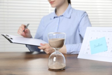 Photo of Deadline. Woman with clipboard and pen at wooden table in office, focus on hourglass