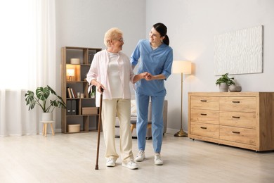 Photo of Nurse assisting senior woman with walking cane indoors. Home health care service