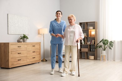 Photo of Nurse assisting senior woman with walking cane indoors. Home health care service