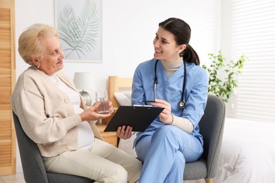 Photo of Nurse with clipboard examining senior woman indoors. Home health care service