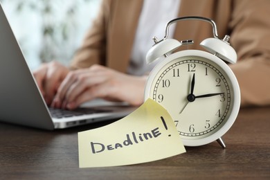 Photo of Deadline. Woman with laptop working at wooden table in office, focus on alarm clock and sticky note