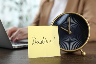 Photo of Deadline. Woman with laptop working at wooden table in office, focus on alarm clock and sticky note
