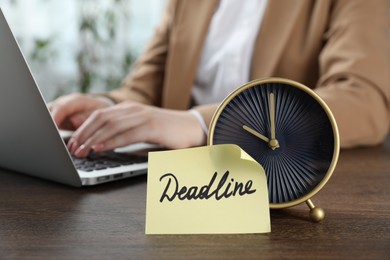 Photo of Deadline. Woman with laptop working at wooden table in office, focus on alarm clock and sticky note