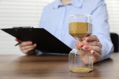 Photo of Deadline. Woman with hourglass and clipboard at wooden table in office, closeup