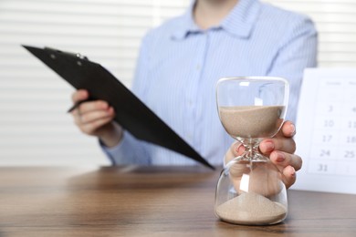 Photo of Deadline. Woman with hourglass and clipboard at wooden table in office, closeup
