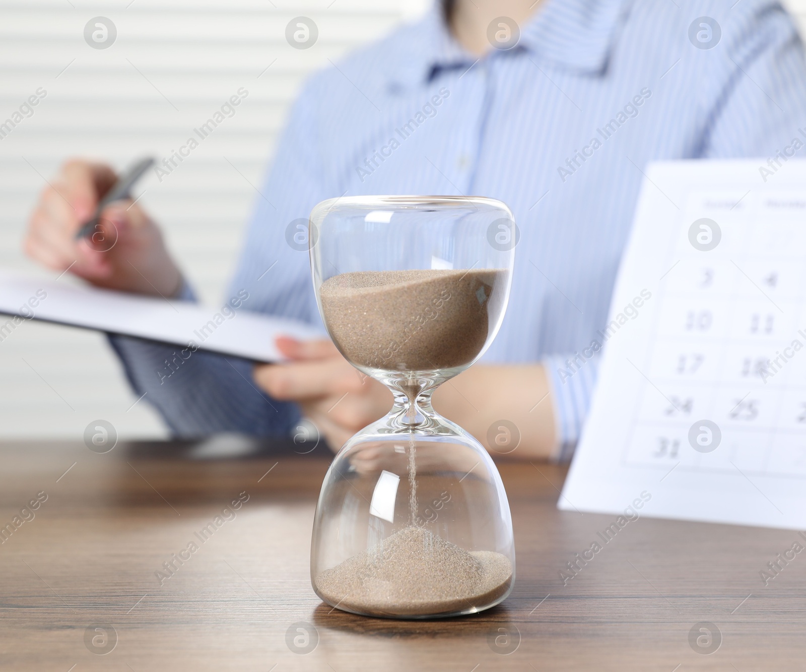 Deadline. Woman with clipboard and pen at wooden table in office, focus on hourglass Photo of Deadline. Woman with clipboard and pen at wooden table in office, focus on hourglass