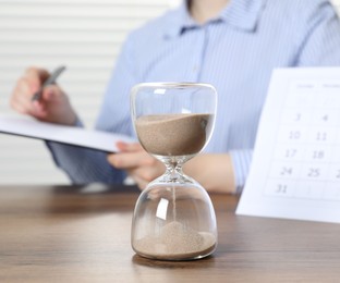 Photo of Deadline. Woman with clipboard and pen at wooden table in office, focus on hourglass