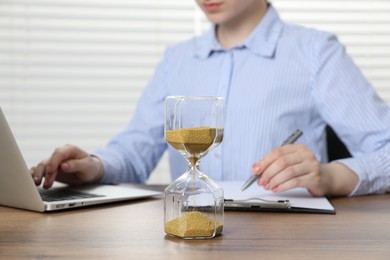 Photo of Deadline. Woman with hourglass and laptop working at wooden table in office, closeup