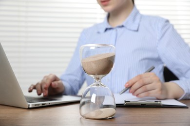 Photo of Deadline. Woman with laptop and clipboard working at wooden table in office, focus on hourglass