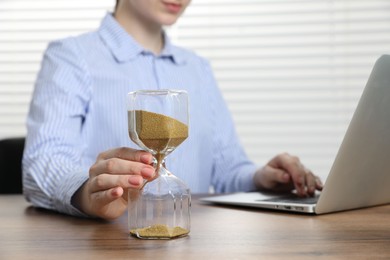 Photo of Deadline. Woman with hourglass and laptop working at wooden table in office, closeup