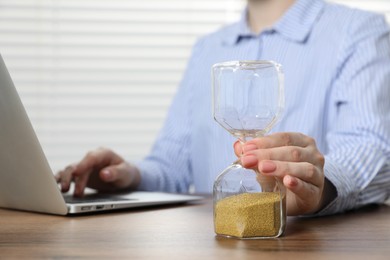 Photo of Deadline. Woman with hourglass and laptop working at wooden table in office, closeup
