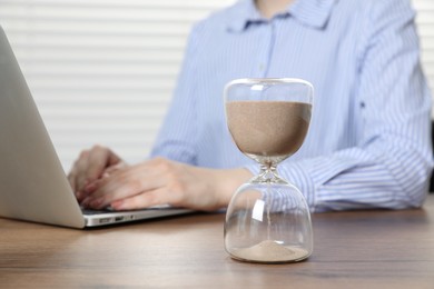 Photo of Deadline. Woman with laptop working at wooden table in office, focus on hourglass