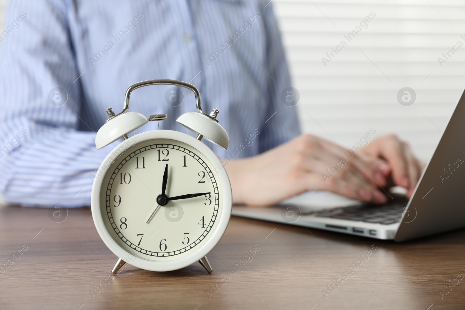 Deadline. Woman with laptop working at wooden table in office, focus on alarm clock Photo of Deadline. Woman with laptop working at wooden table in office, focus on alarm clock