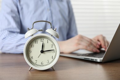 Photo of Deadline. Woman with laptop working at wooden table in office, focus on alarm clock