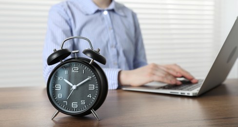 Photo of Deadline. Woman with laptop working at wooden table in office, focus on alarm clock