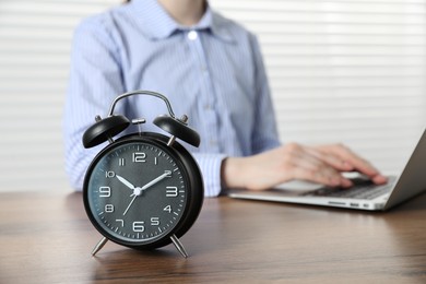 Photo of Deadline. Woman with laptop working at wooden table in office, focus on alarm clock