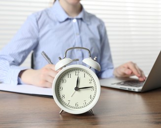 Photo of Deadline. Woman with laptop and clipboard working at wooden table in office, focus on alarm clock