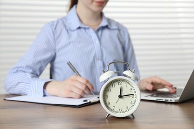 Photo of Deadline. Woman with laptop and clipboard working at wooden table in office, focus on alarm clock