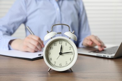 Photo of Deadline. Woman with laptop and clipboard working at wooden table in office, focus on alarm clock