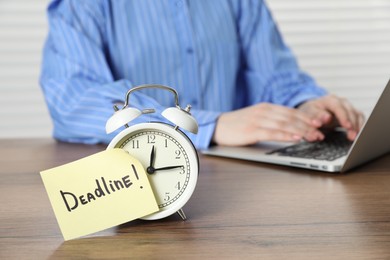 Photo of Deadline. Woman with laptop working at wooden table in office, focus on alarm clock and sticky note