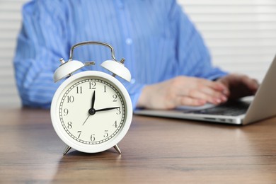 Photo of Deadline. Woman with laptop working at wooden table in office, focus on alarm clock