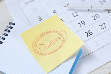 Photo of Sticky note with word Deadline, notebook, pen and calendar on table, closeup