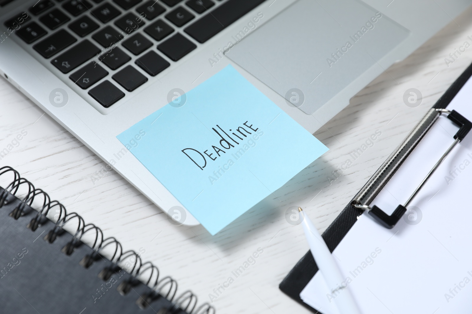 Photo of Sticky note with word Deadline, laptop, notebook, clipboard and pen on white wooden table, closeup