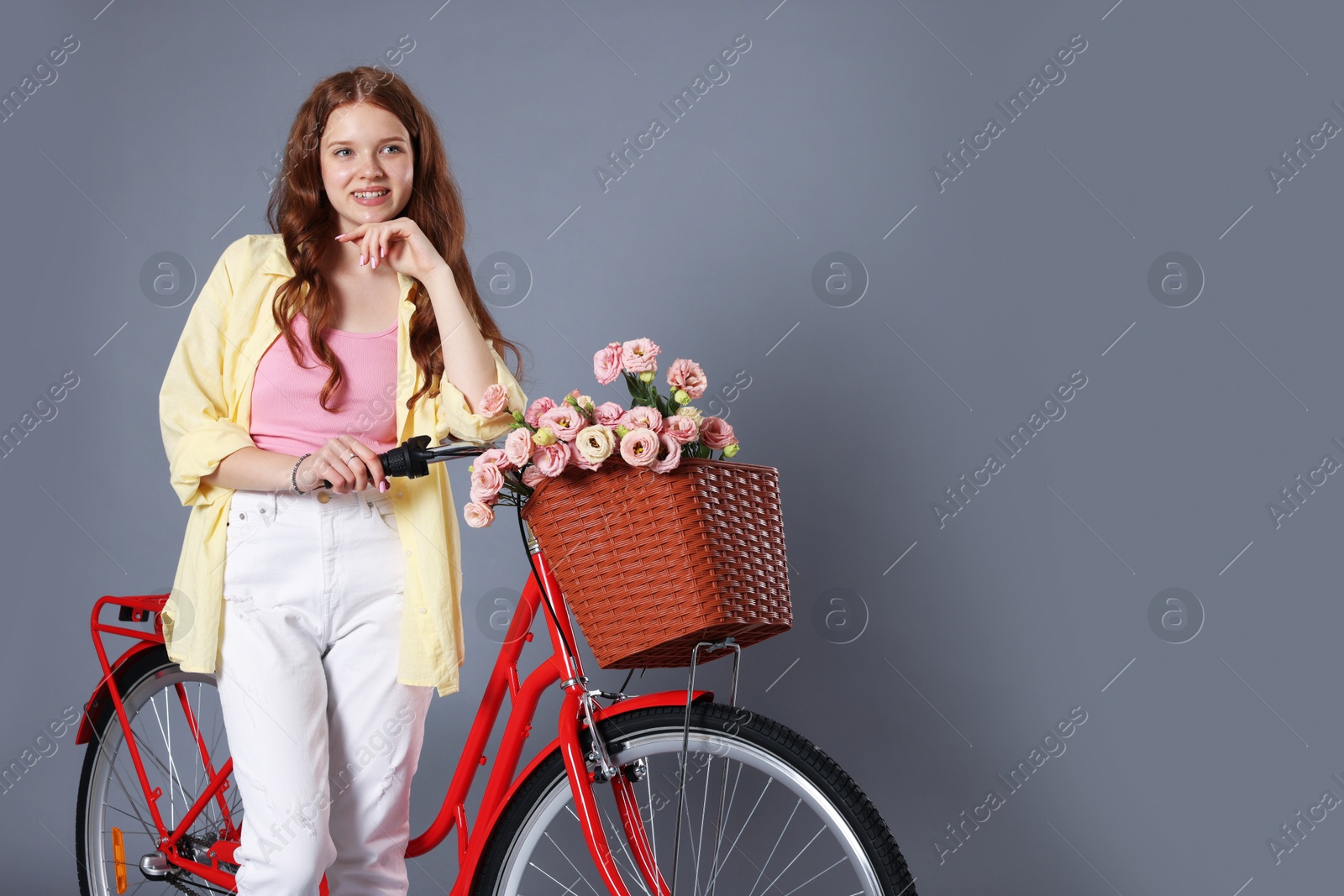 Smiling teenage girl and bicycle with basket of flowers against grey background. Space for text Photo of Smiling teenage girl and bicycle with basket of flowers against grey background. Space for text