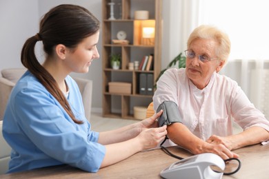 Photo of Nurse measuring patient's blood pressure at wooden table indoors. Home health care service