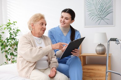 Photo of Nurse with clipboard examining senior woman indoors. Home health care service