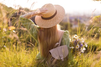 Photo of Woman with beautiful wildflowers, hat and backpack in meadow on sunny day, back view