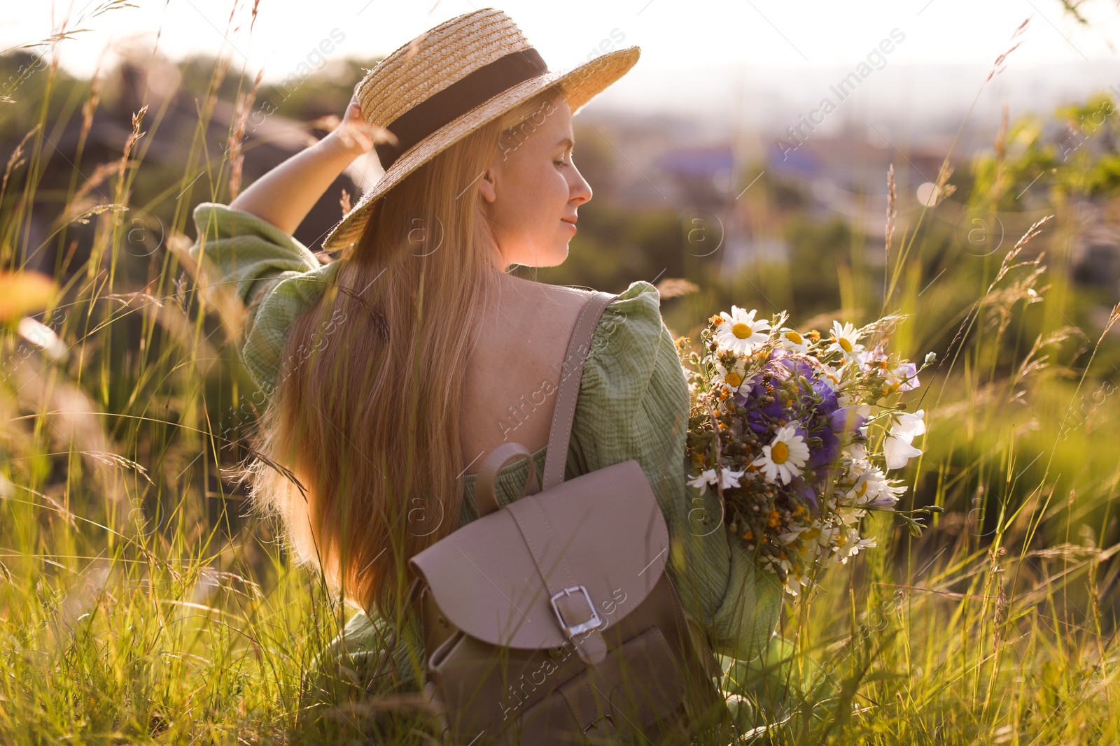 Woman with beautiful wildflowers, hat and backpack in meadow on sunny day, back view Photo of Woman with beautiful wildflowers, hat and backpack in meadow on sunny day, back view