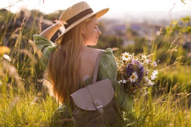 Photo of Woman with beautiful wildflowers, hat and backpack in meadow on sunny day, back view