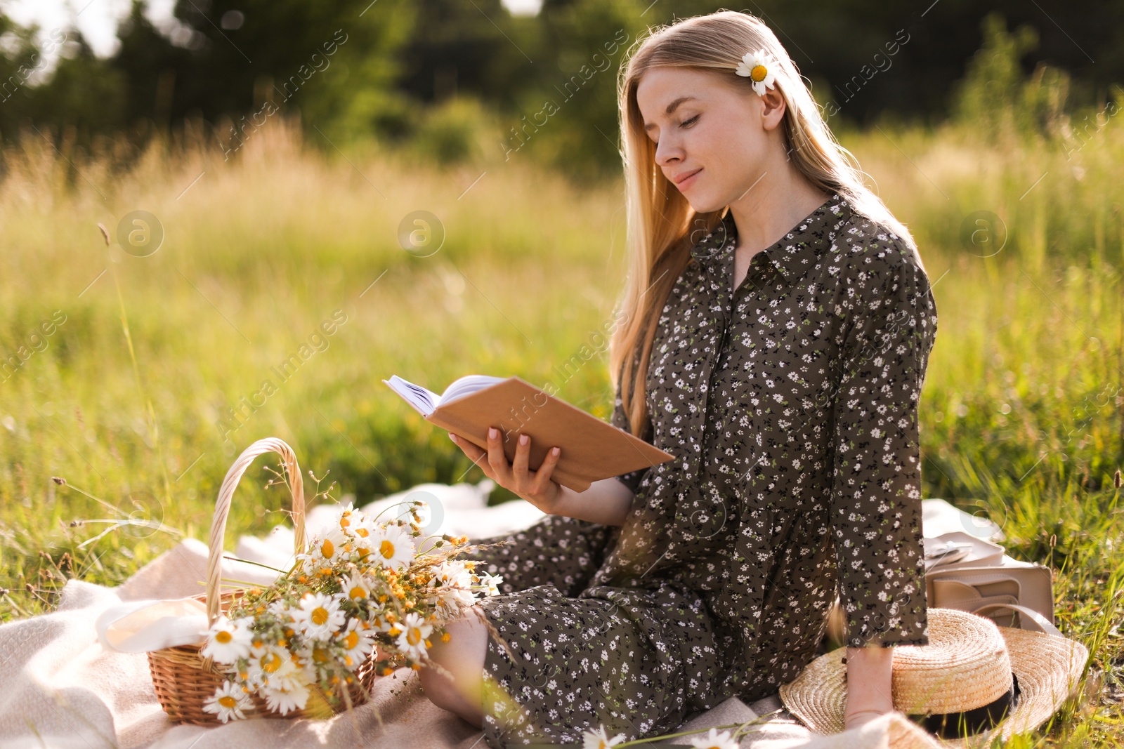 Photo of Beautiful woman with bouquet of wildflowers reading book on blanket in meadow