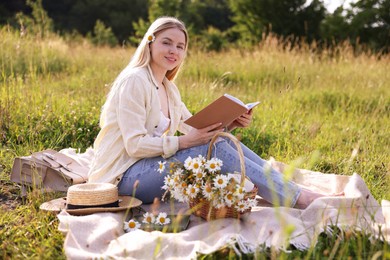 Photo of Woman with bouquet of wildflowers reading book on blanket in meadow