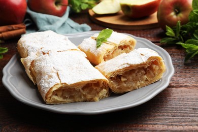 Photo of Pieces of tasty apple strudel with powdered sugar, mint and fruits on wooden table, closeup