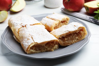 Photo of Pieces of tasty apple strudel with powdered sugar and fruits on white marble table, closeup