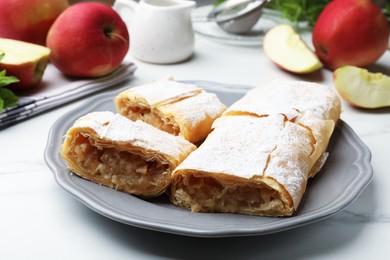 Photo of Pieces of tasty apple strudel with powdered sugar and fruits on white marble table, closeup