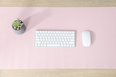 Photo of Computer keyboard, mouse, houseplant and pink mousepad on wooden table at workplace, flat lay