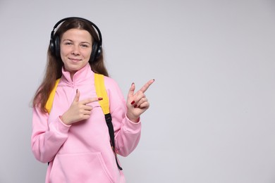 Photo of Teenage girl in headphones with backpack pointing at something on light grey background, space for text