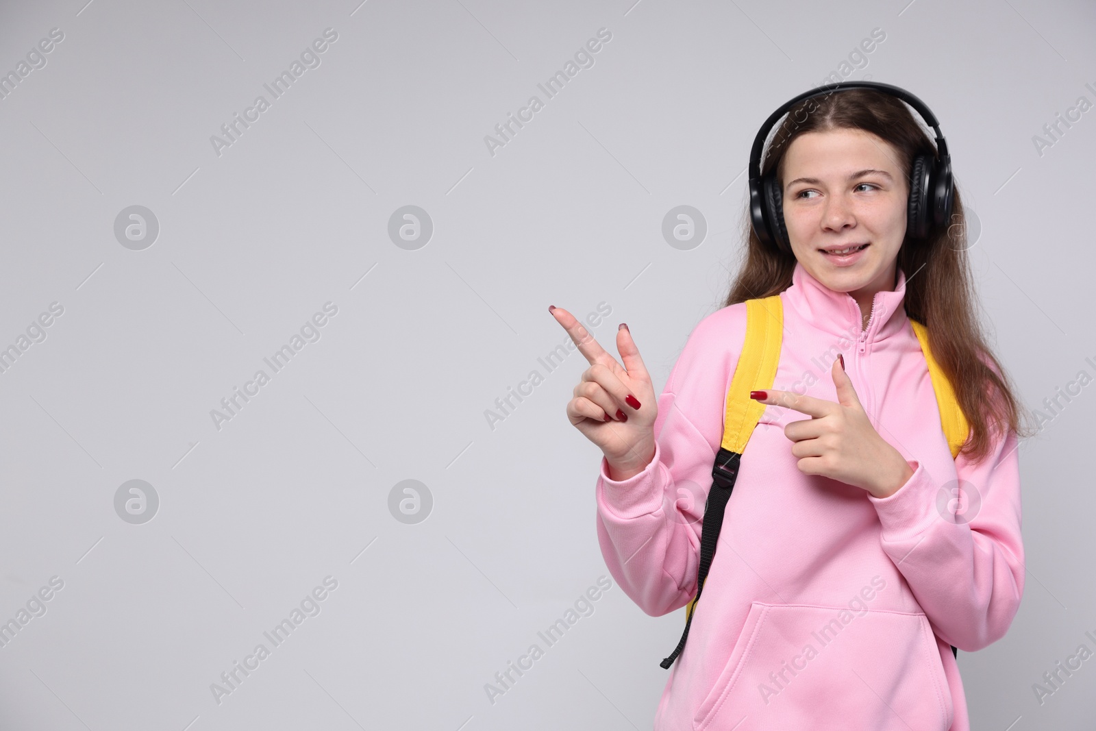 Teenage girl in headphones with backpack pointing at something on light grey background, space for text Photo of Teenage girl in headphones with backpack pointing at something on light grey background, space for text
