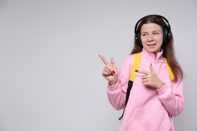 Photo of Teenage girl in headphones with backpack pointing at something on light grey background, space for text