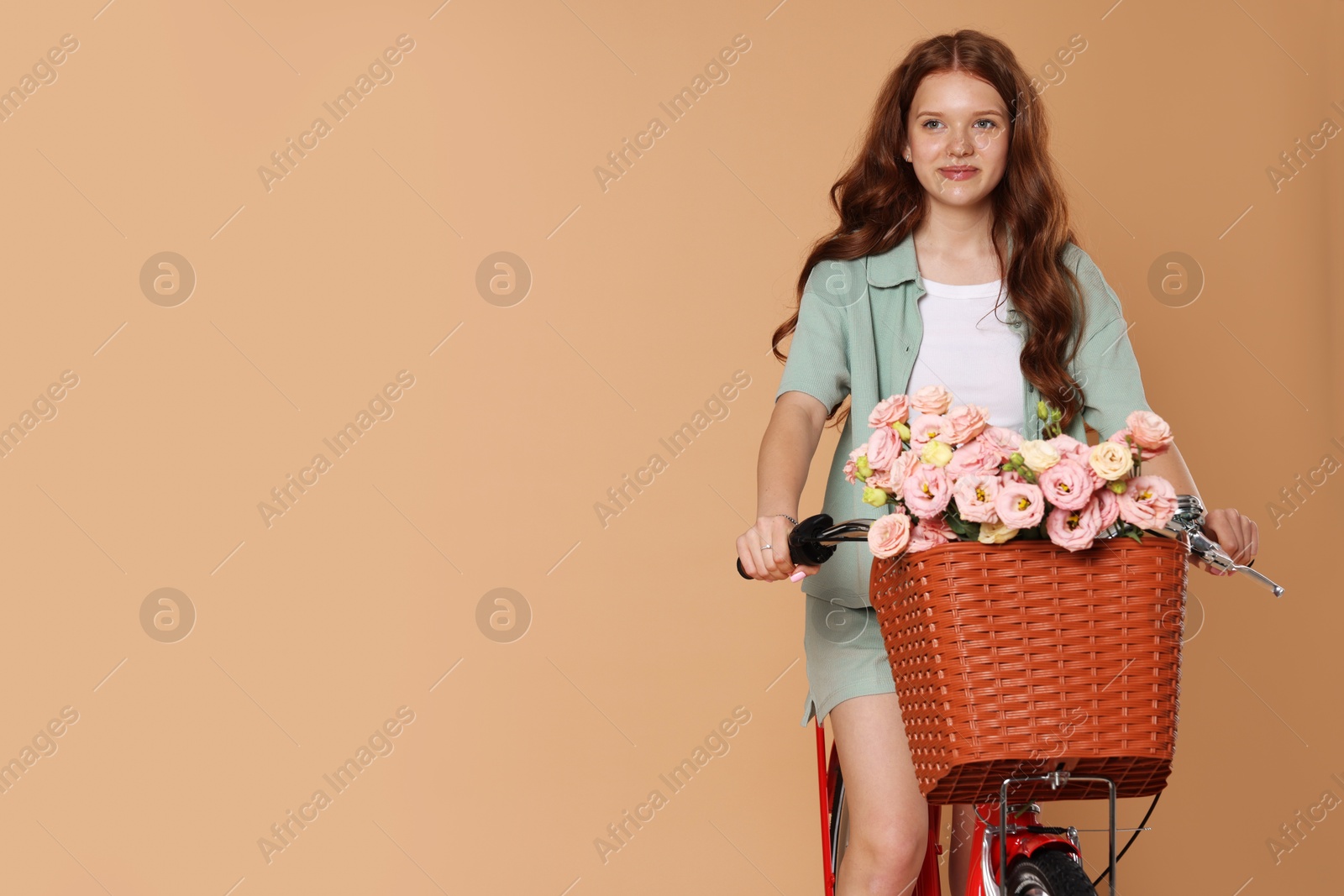 Beautiful teenage girl riding bicycle with basket of flowers against beige background. Space for text Photo of Beautiful teenage girl riding bicycle with basket of flowers against beige background. Space for text