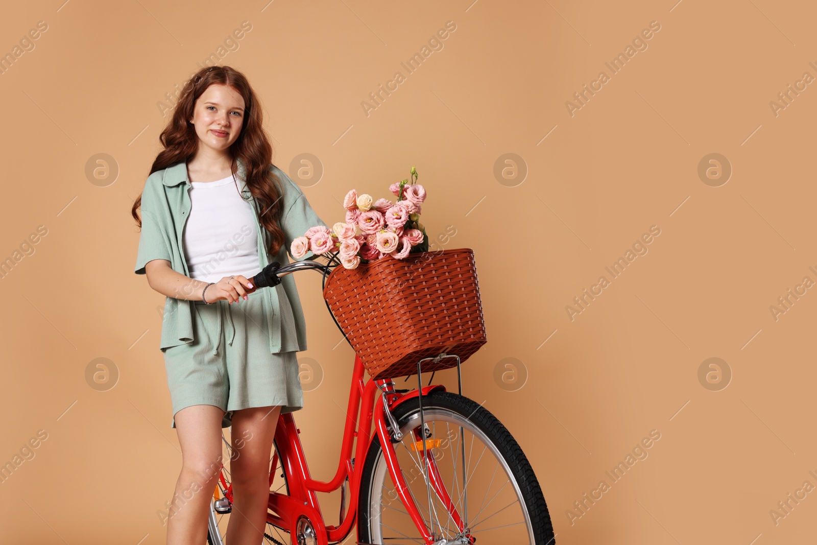Beautiful teenage girl and bicycle with basket of flowers against beige background. Space for text Photo of Beautiful teenage girl and bicycle with basket of flowers against beige background. Space for text