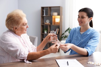Photo of Nurse giving glass of water and pills to senior woman at wooden table indoors. Home health care service