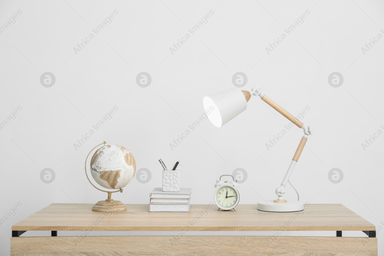 Lamp, books, stationery, alarm clock and globe on wooden table near white wall indoors Photo of Lamp, books, stationery, alarm clock and globe on wooden table near white wall indoors