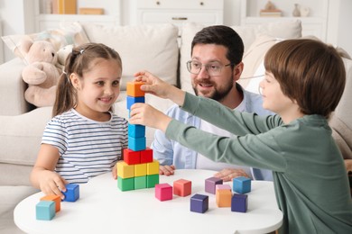 Photo of Children and their father building tower with colorful cubes at white table indoors