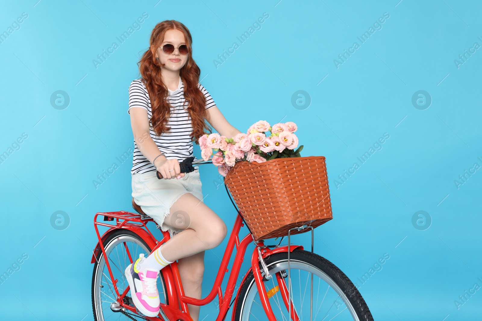 Beautiful teenage girl in sunglasses riding bicycle with basket of flowers against light blue background Photo of Beautiful teenage girl in sunglasses riding bicycle with basket of flowers against light blue background