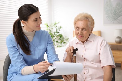 Photo of Nurse with clipboard examining senior woman indoors. Home health care service